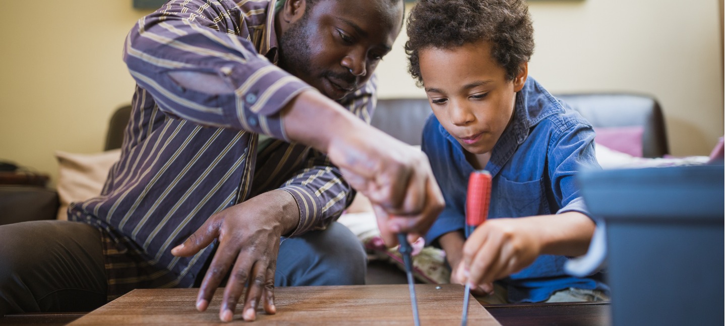 A dad and son using screw drivers on a piece of furniture.