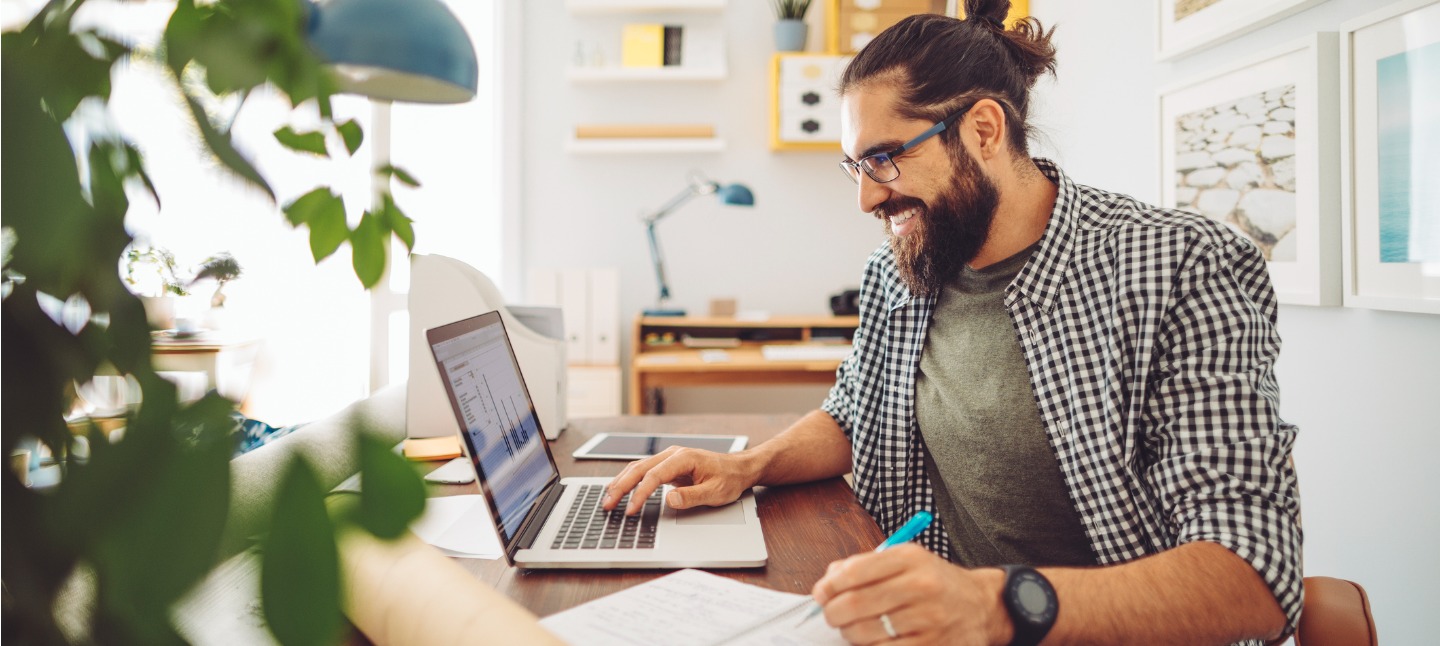 Young man with a beard at home, smiling while budgeting on his laptop.